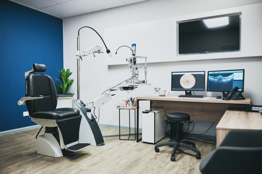 An eye exam room with a blue wall, exam chair, slit lamp, plant and two screens, no lens